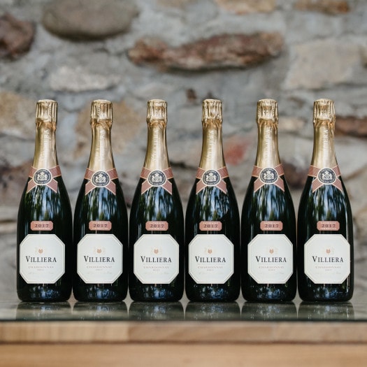 Six bottles of Villiera Brut Villiera Brut Natural 2021 sparkling wine against a stone wall.